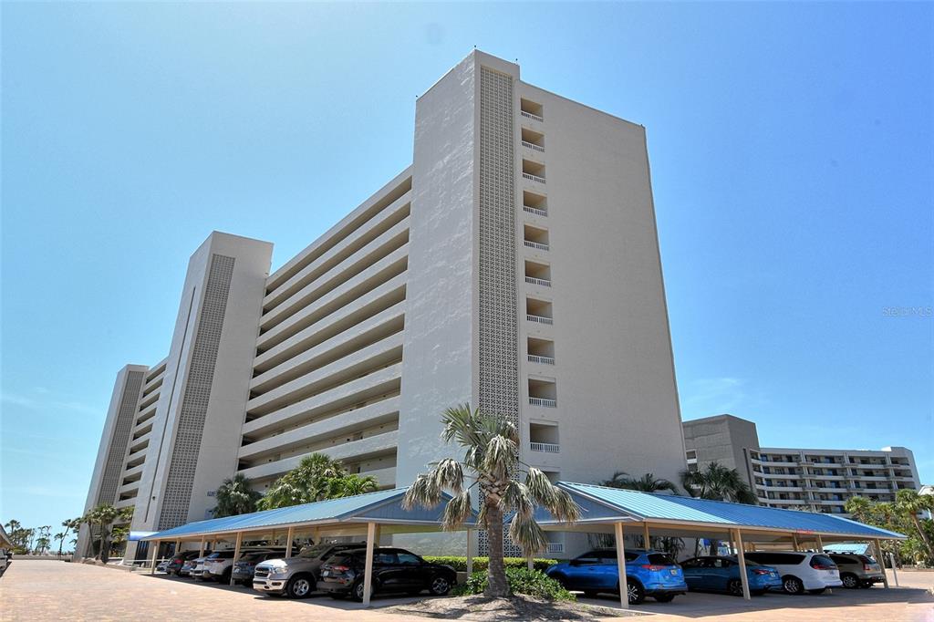 A tall, beige multi-story apartment building stands behind a covered parking area with several cars — 5920 MIDNIGHT PASS ROAD UNIT 406, SARASOTA, FL