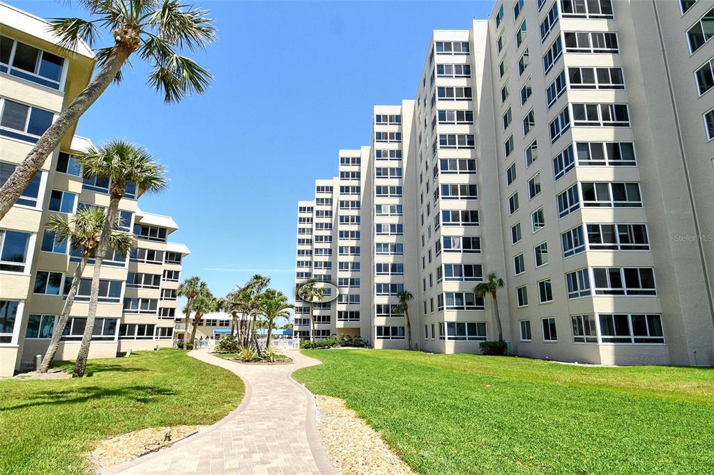 A paved walkway leads through a grassy courtyard between two large, multi-story residential buildings — 5920 MIDNIGHT PASS ROAD UNIT 406, SARASOTA, FL