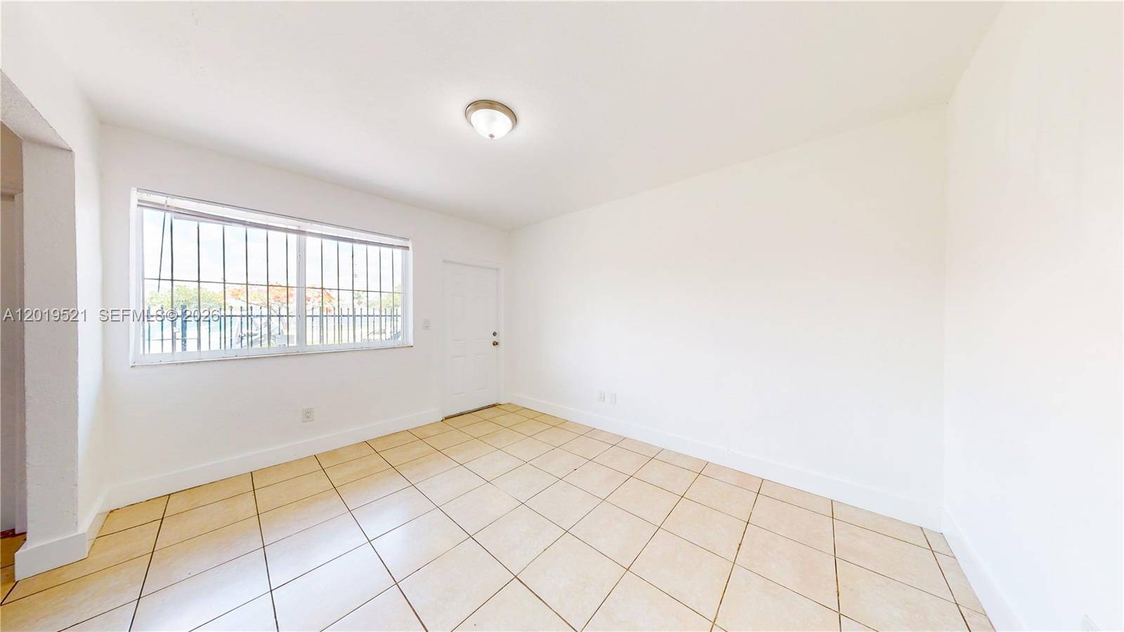 An empty room features white walls, beige tile flooring, a window with security bars, and a door — 1190 NW 65th St # 2, Miami FL 33150, MIAMI, FL