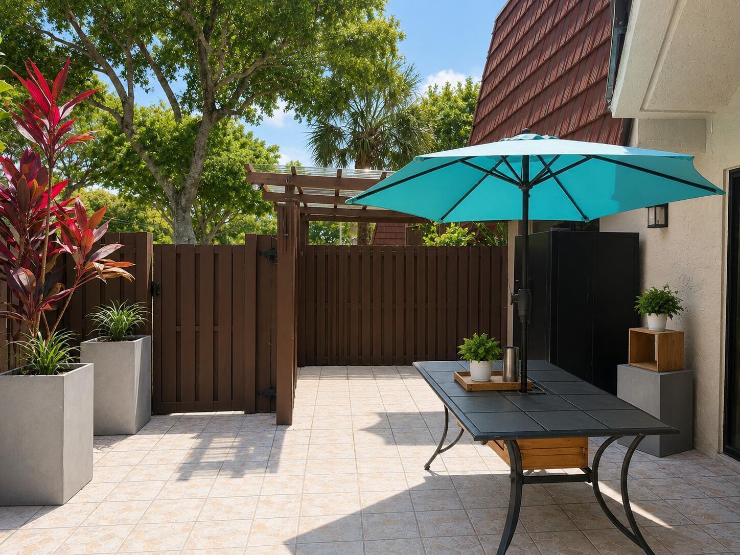 A patio features a tiled floor, a turquoise umbrella over a table, and wooden fencing — 8259 Boca Rio, Boca Raton FL 33433, BOCA RATON, FL