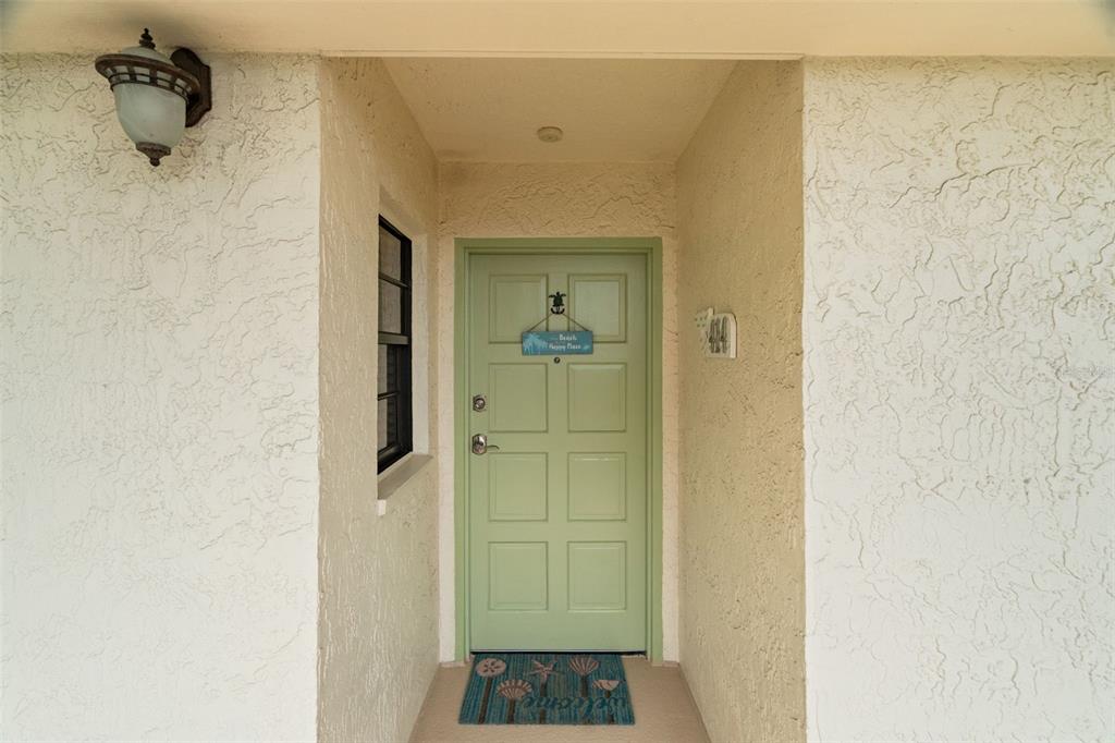 A light green front door with a welcome mat sits under a porch light — 19531 GULF BOULEVARD UNIT 414, INDIAN SHORES, FL