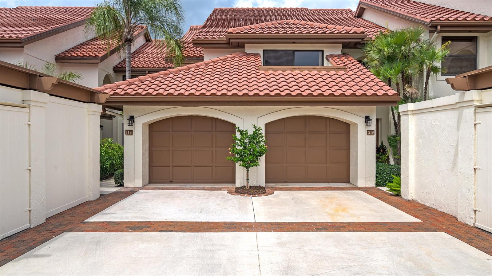 A two-car garage with brown doors and a small tree sits beneath a tiled roof — 16695 Traders Crossing N # 118, Jupiter FL 33477, JUPITER, FL