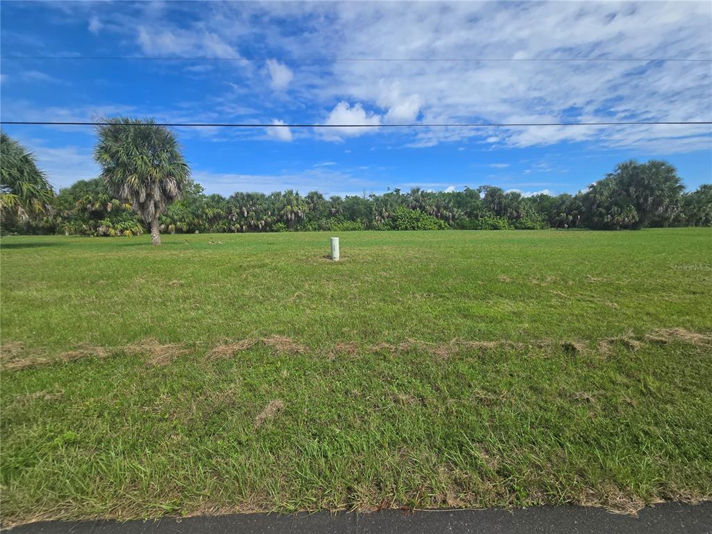 A flat, grassy vacant lot with a utility pipe and trees in the background — 16947 ACAPULCO ROAD, PUNTA GORDA, FL