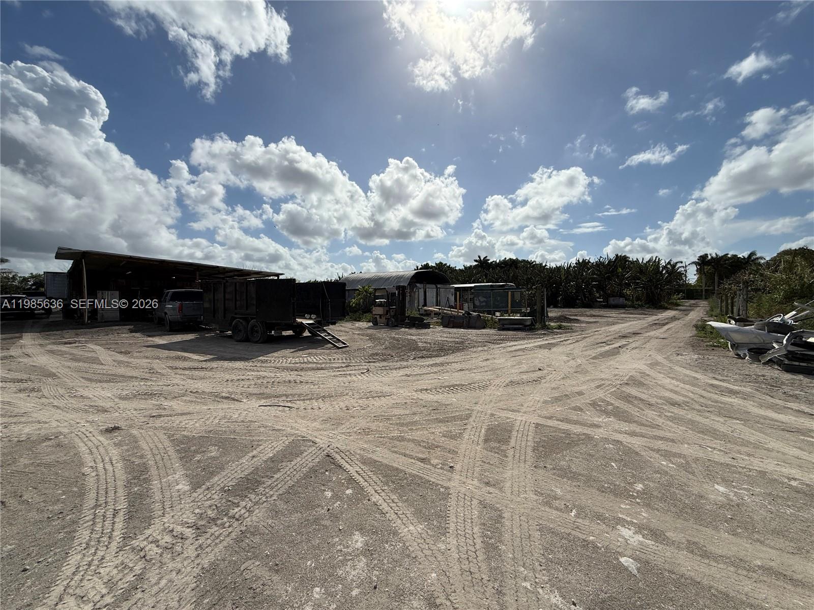 A dirt lot features a large open-sided shed, a trailer, and scattered equipment under clouds — 16455 SW 207, Miami FL 33187, MIAMI, FL