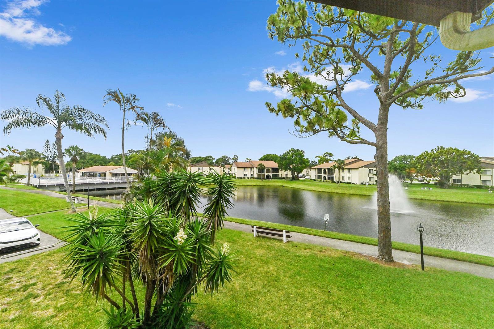 A view of a pond, fountain, and apartment buildings from a grassy lawn with trees — 3561 Pine Tree Court # 2, FL 33463, FL