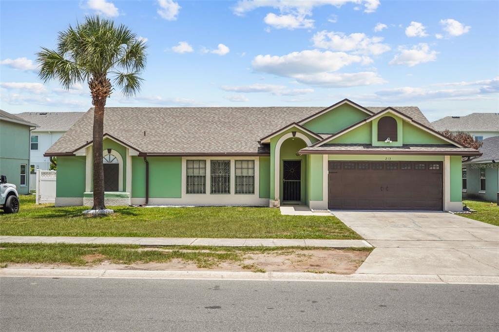 A single-story house with green stucco walls, a brown garage door, and a palm tree — 213 COMPETITION DRIVE, KISSIMMEE, FL