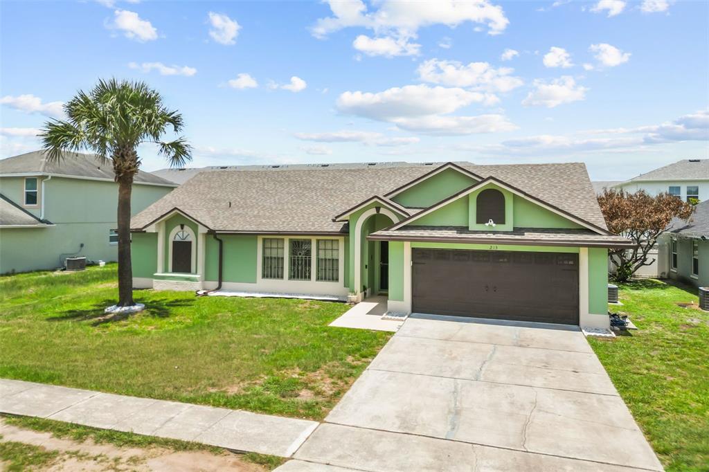 A single-story green house with a brown roof, a two-car garage, and a palm tree — 213 COMPETITION DRIVE, KISSIMMEE, FL