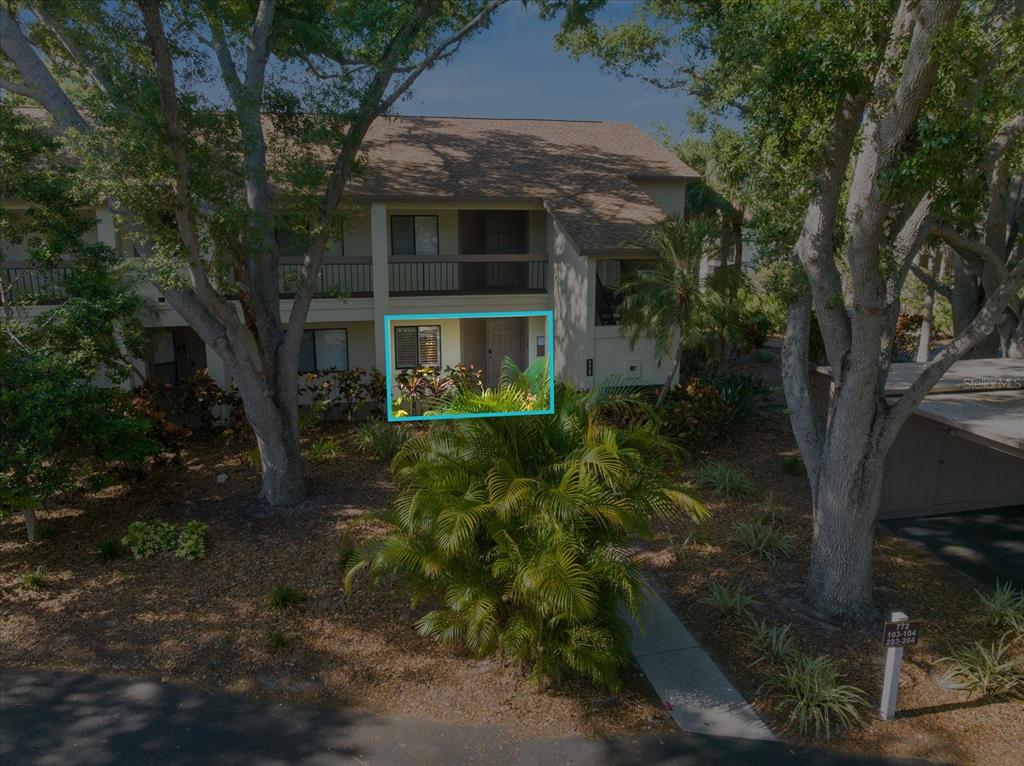An exterior view of a ground-floor apartment entrance surrounded by trees and tropical landscaping — 772 BIRD BAY DRIVE N UNIT 104, VENICE, FL