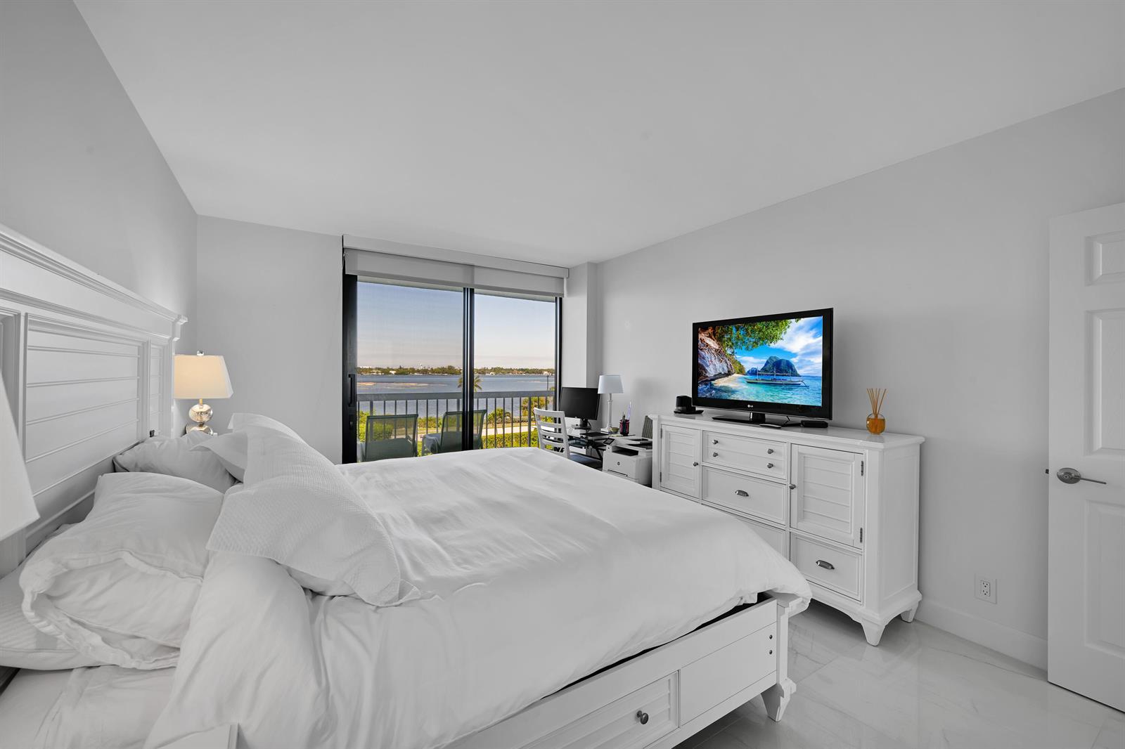 A white bedroom features a bed, a dresser with a television, and a balcony view — 3300 S Ocean Boulevard # 306n, Palm Beach FL 33480, PALM BEACH, FL