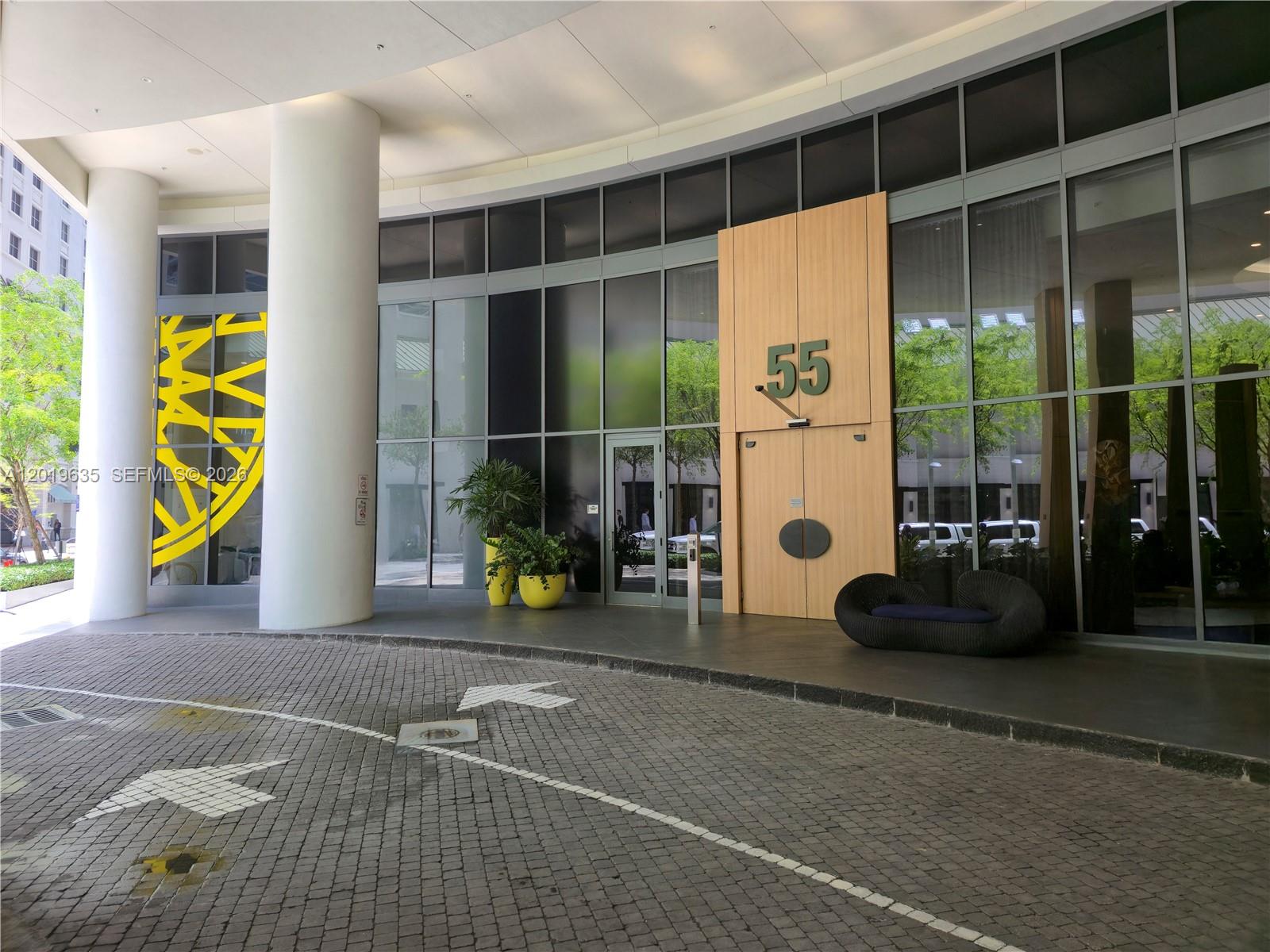 The exterior entrance of a building features a curved driveway, glass facade, and wooden doors — 55 SW 9th St # 1810, Miami FL 33130, MIAMI, FL