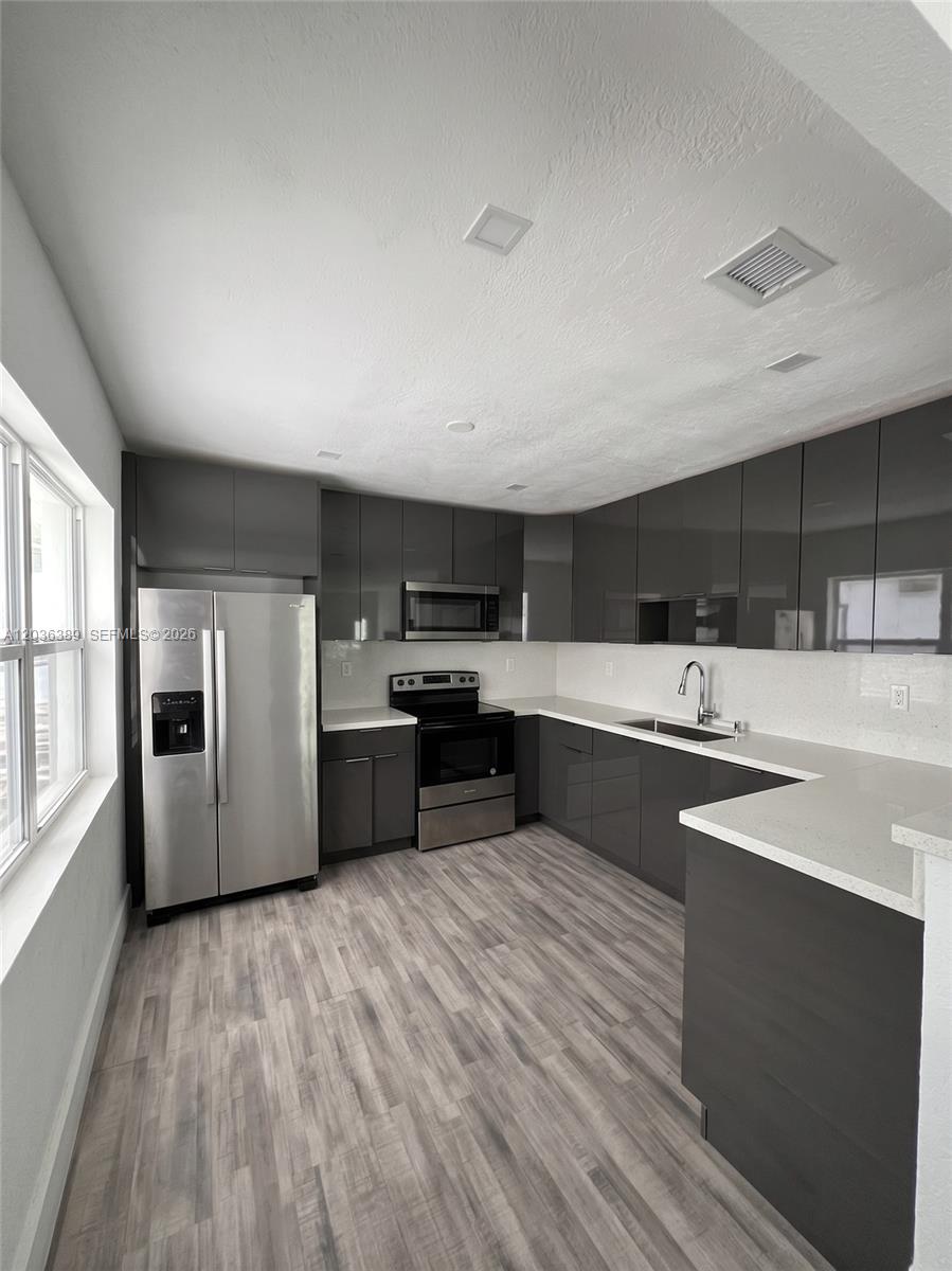 A modern kitchen features dark gray high-gloss cabinets, stainless steel appliances, and light wood flooring — 2847 Polk St # 1, Hollywood FL 33020, HOLLYWOOD, FL