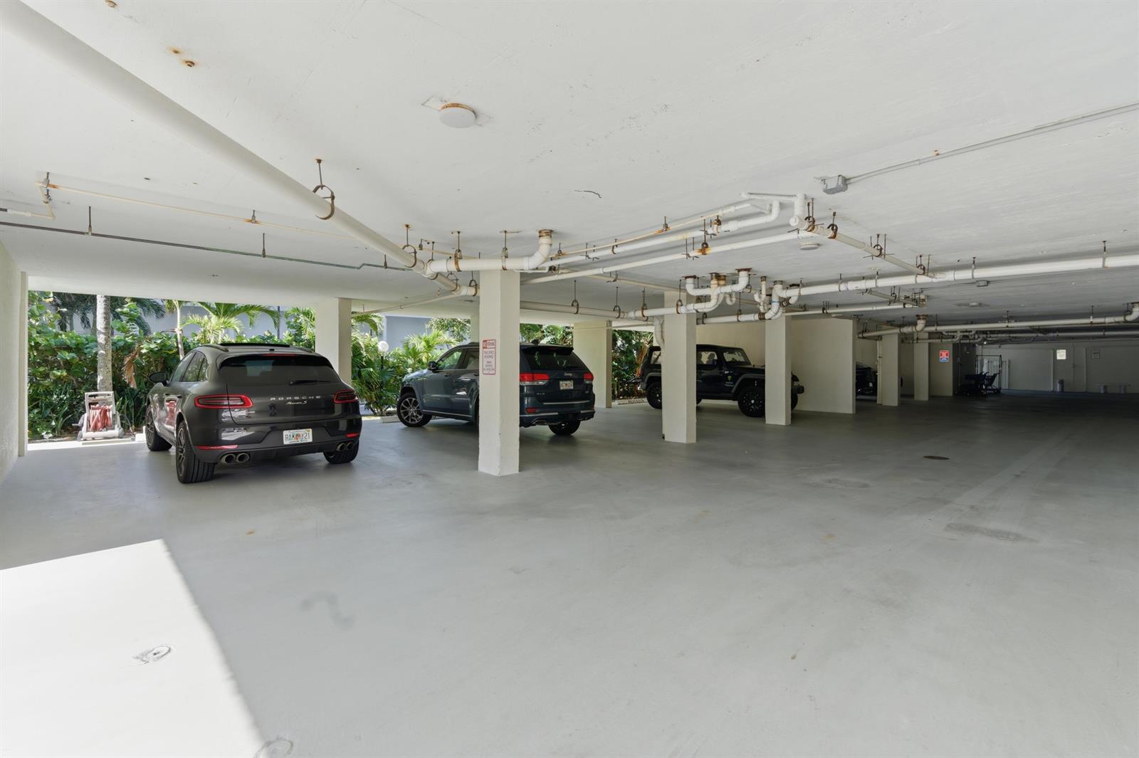 A covered parking garage features concrete pillars, exposed overhead pipes, and several parked vehicles — 3581 S Ocean Boulevard Apt 4e, South Palm Beach FL 33480, SOUTH PALM BEACH, FL