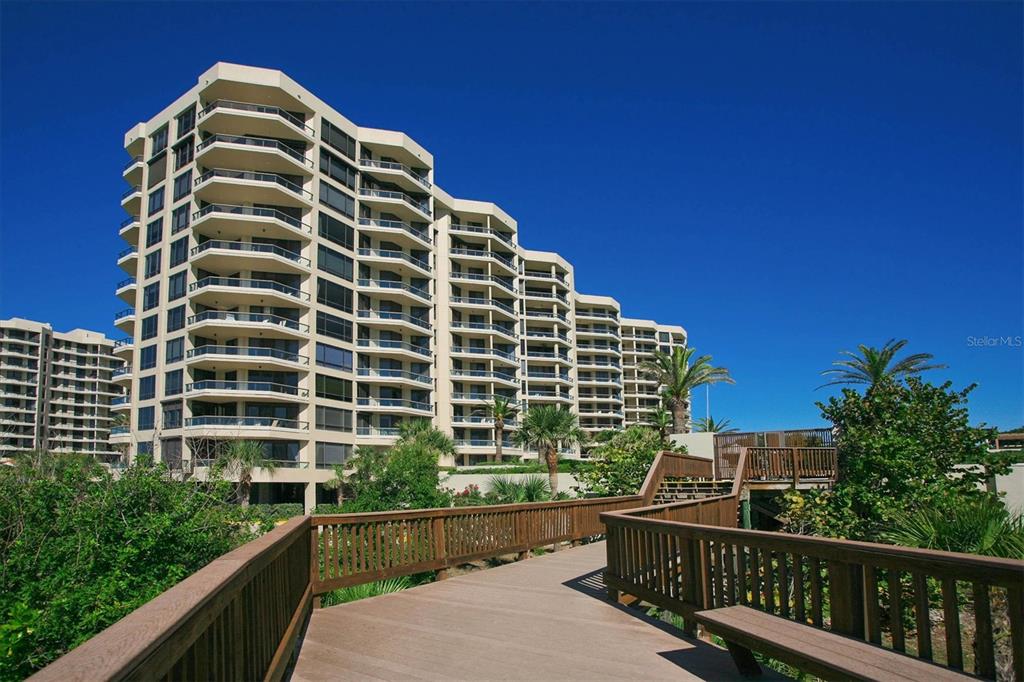 A wooden boardwalk leads toward a multi-story residential building with balconies under a clear sky — 1211 GULF OF MEXICO DRIVE UNIT 405, LONGBOAT KEY, FL