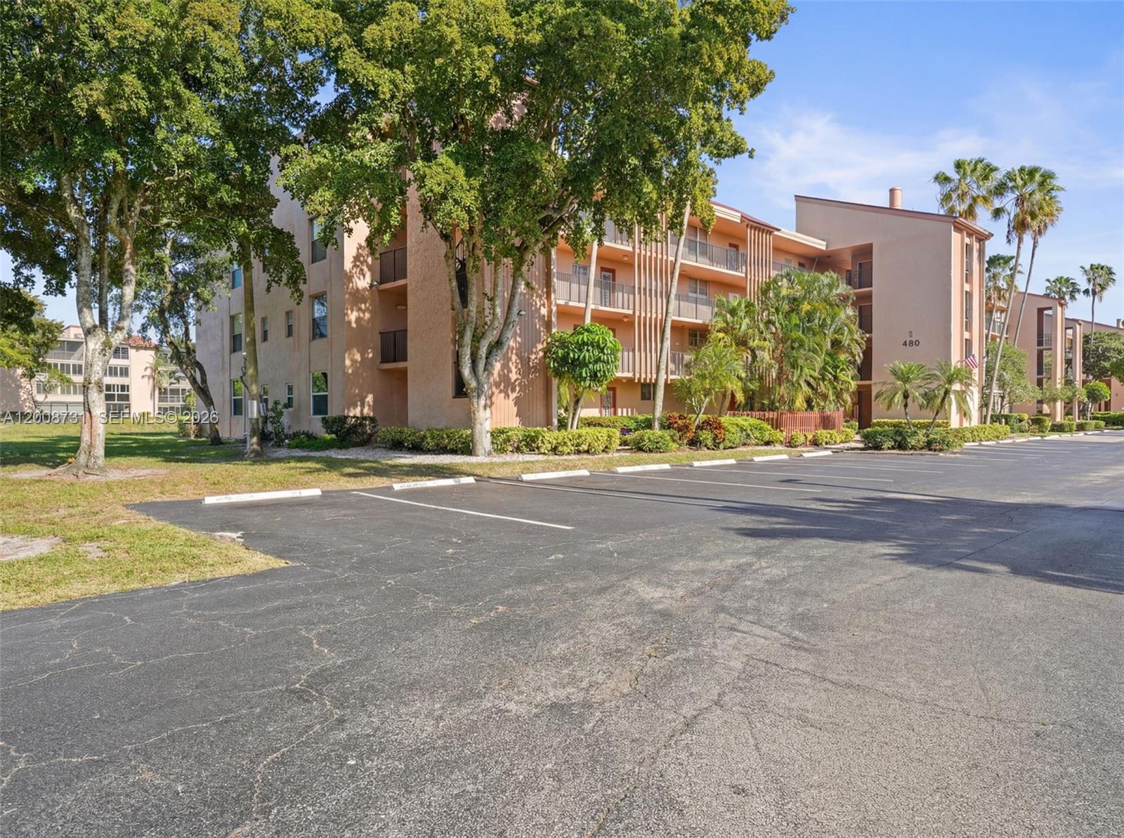 A multi-story apartment building with balconies stands behind a paved parking lot and trees — 480 NW 76th Ave # 102, Margate FL 33063, MARGATE, FL