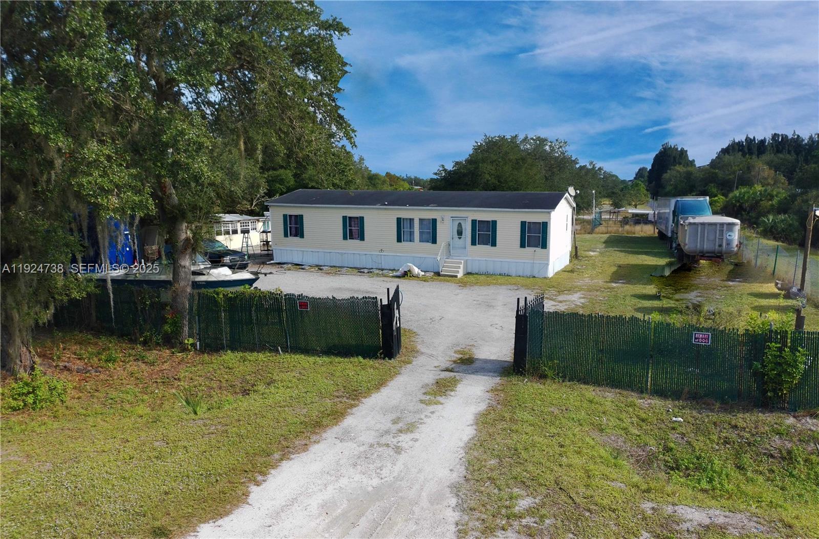 An exterior view of a single-story manufactured home with a gravel driveway and fenced yard — 225 N Estribo ST, Clewiston FL 33440, CLEWISTON, FL
