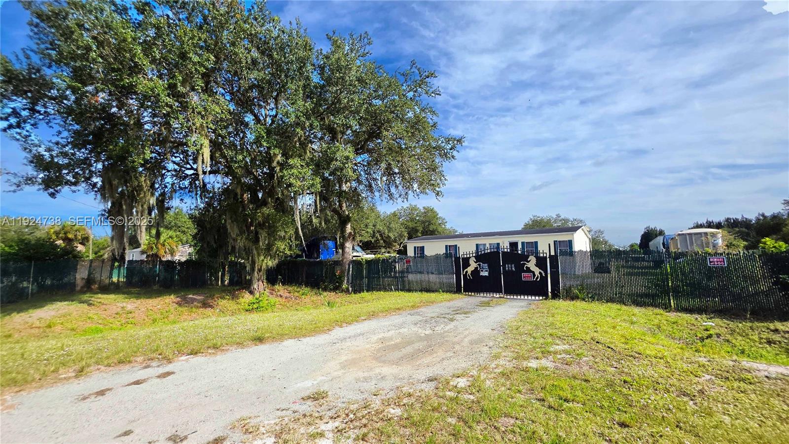 A gravel driveway leads to a fenced property featuring a house and large oak trees — 225 N Estribo ST, Clewiston FL 33440, CLEWISTON, FL