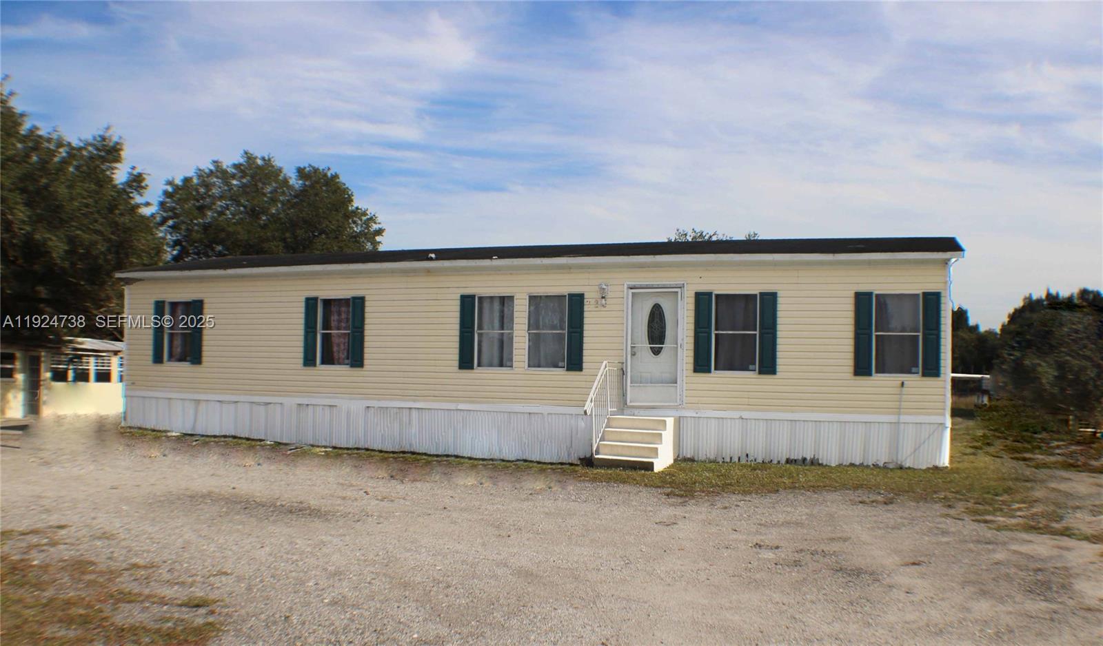 A single-story mobile home with yellow siding and dark shutters sits on a gravel lot — 225 N Estribo ST, Clewiston FL 33440, CLEWISTON, FL