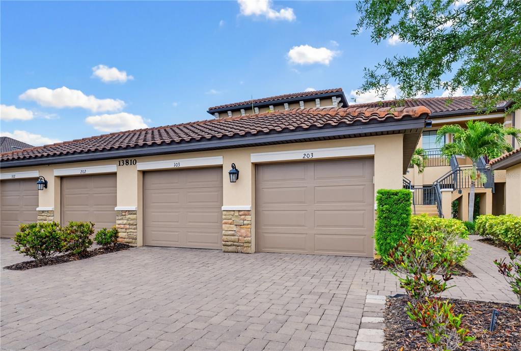 A row of three tan garage doors with stone accents under a tiled roof — 13810 MESSINA LOOP UNIT 203, BRADENTON, FL