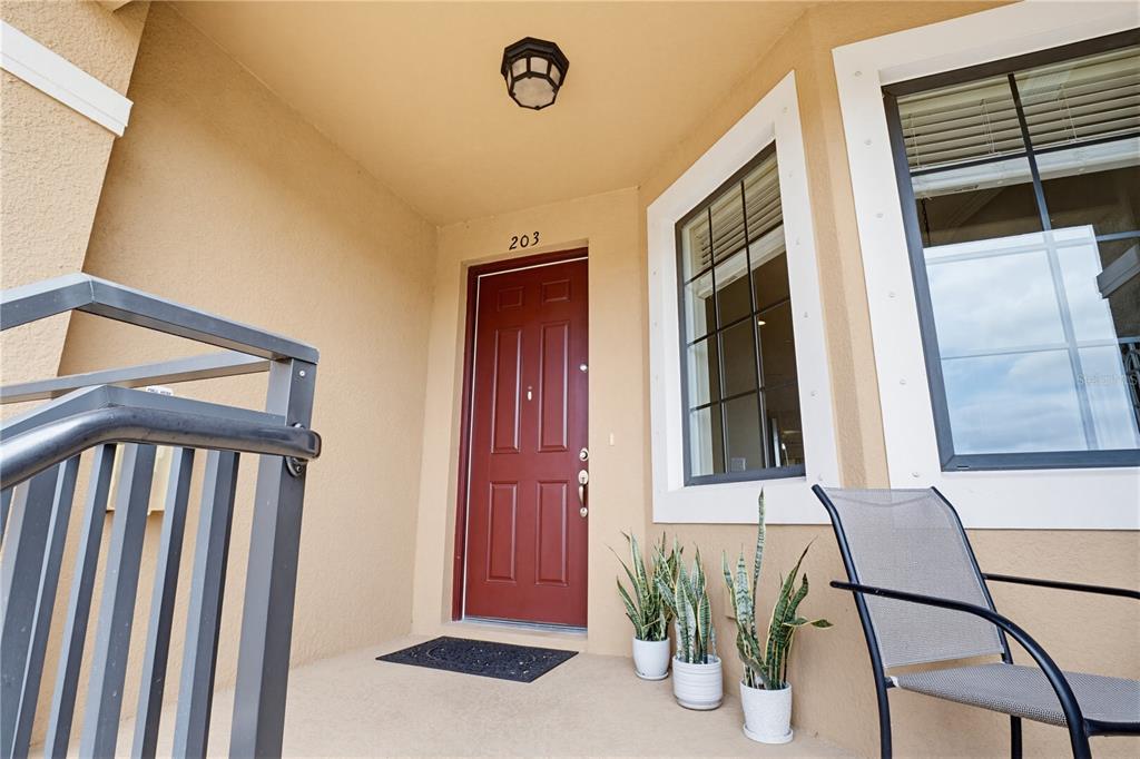 A red front door with the number 203 sits on a porch with potted plants — 13810 MESSINA LOOP UNIT 203, BRADENTON, FL