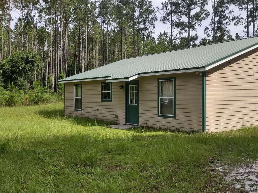 A tan, single-story house with a green metal roof sits in a grassy, wooded area — 4651 NE 108TH PLACE, CHIEFLAND, FL
