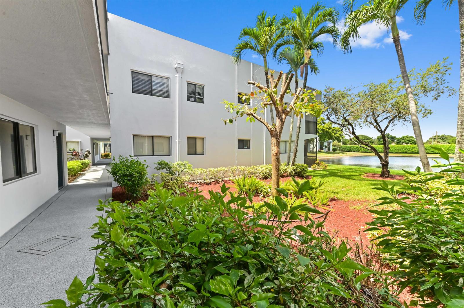 A white multi-story apartment building exterior with a walkway, landscaping, and a nearby pond — 15054 Ashland Way # 84, Delray Beach FL 33484, DELRAY BEACH, FL