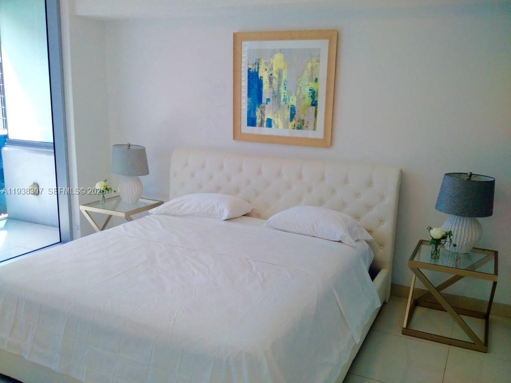 A bedroom features a tufted white headboard, two glass-topped side tables, and abstract wall art — 200 Biscayne Boulevard Way # 906, Miami FL 33131, MIAMI, FL