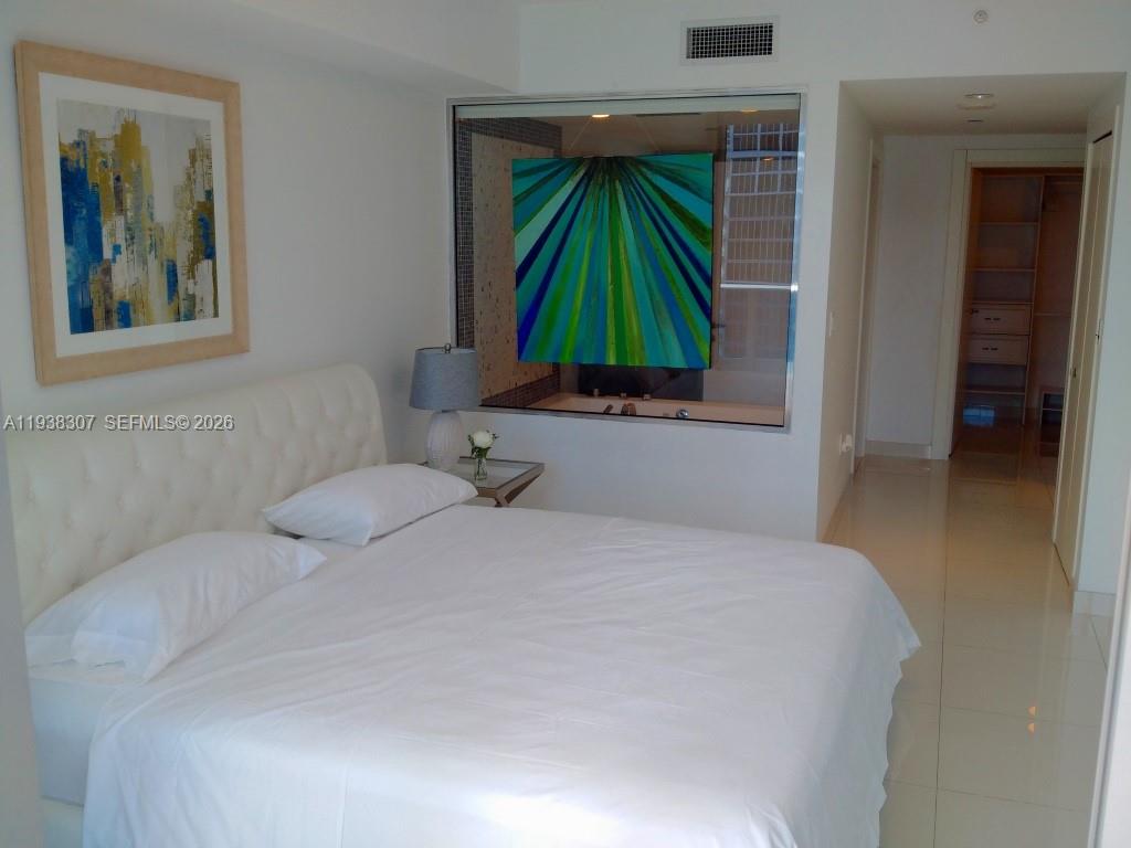 A bedroom features a white tufted headboard, a glass wall partition, and tile flooring — 200 Biscayne Boulevard Way # 906, Miami FL 33131, MIAMI, FL