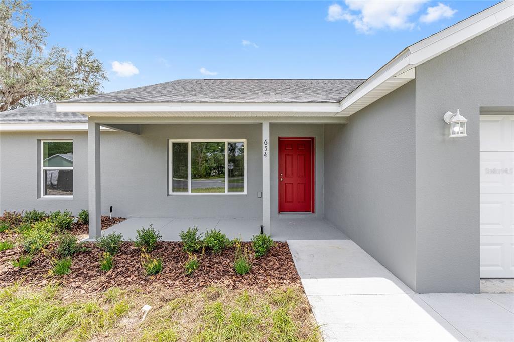A gray house exterior features a red front door, a covered porch, and landscaping — 19788 SW FLAMINGO DRIVE, DUNNELLON, FL