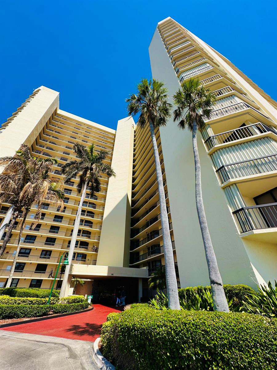 A low-angle view of a tall, light-colored apartment building with balconies and palm trees — 9500 S Ocean Drive # 108, Jensen Beach FL 34957, JENSEN BEACH, FL