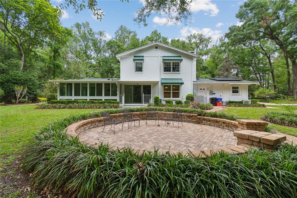 A white two-story house features a circular brick patio with seating in a wooded yard — 2006 NW 23RD STREET, GAINESVILLE, FL