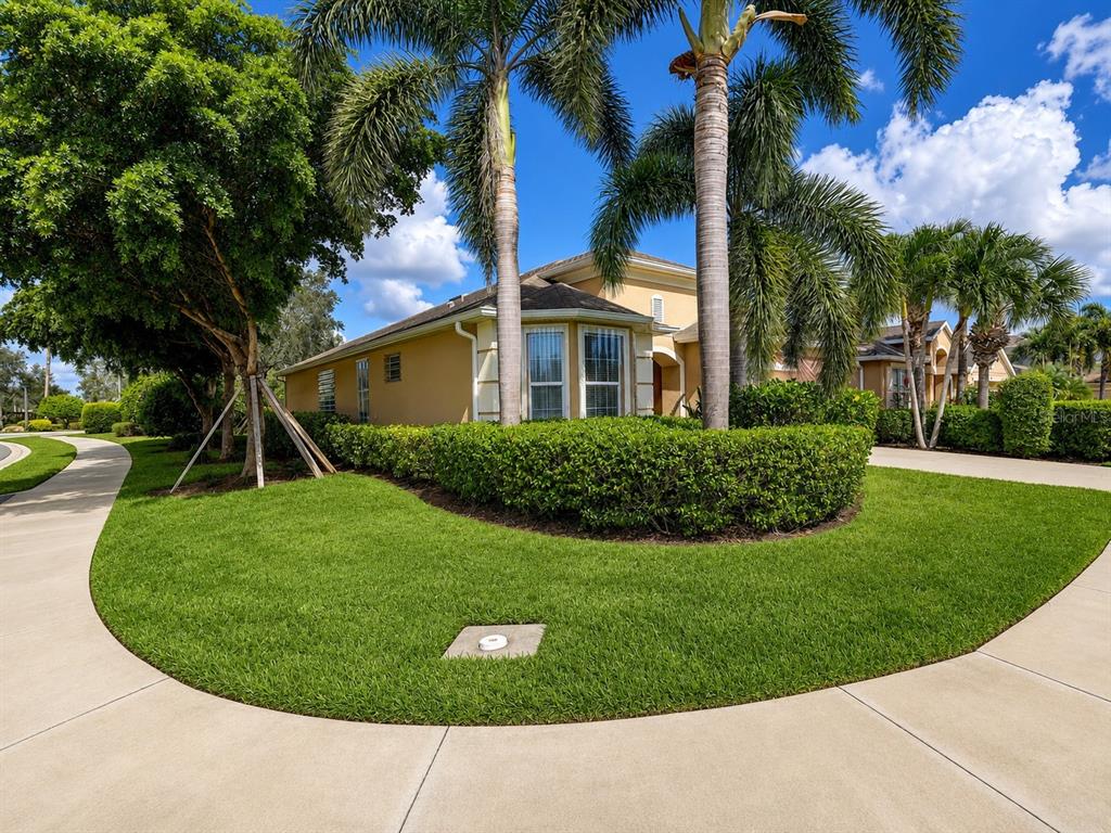 A tan house with a manicured hedge and palm trees sits behind a curved sidewalk — 2542 CORTENOVA COURT, VENICE, FL
