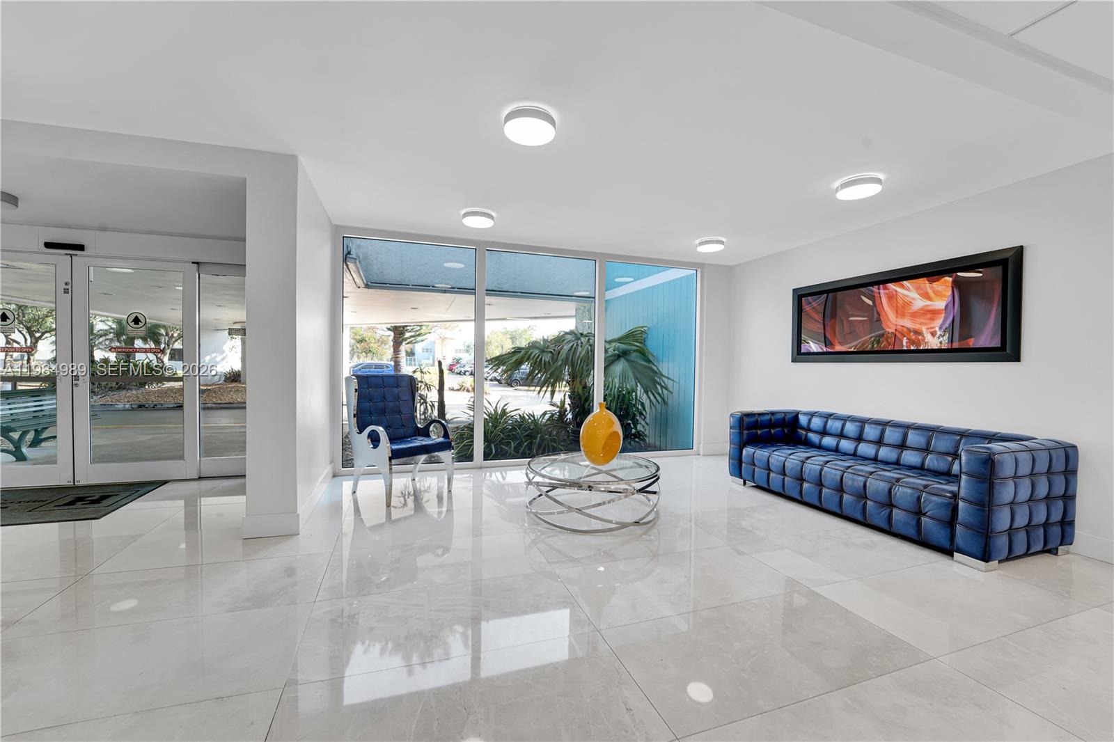 A modern lobby features a blue tufted sofa, matching armchair, glass coffee table, and tile flooring — 919 Hillcrest Dr # 210, Hollywood FL 33021, HOLLYWOOD, FL