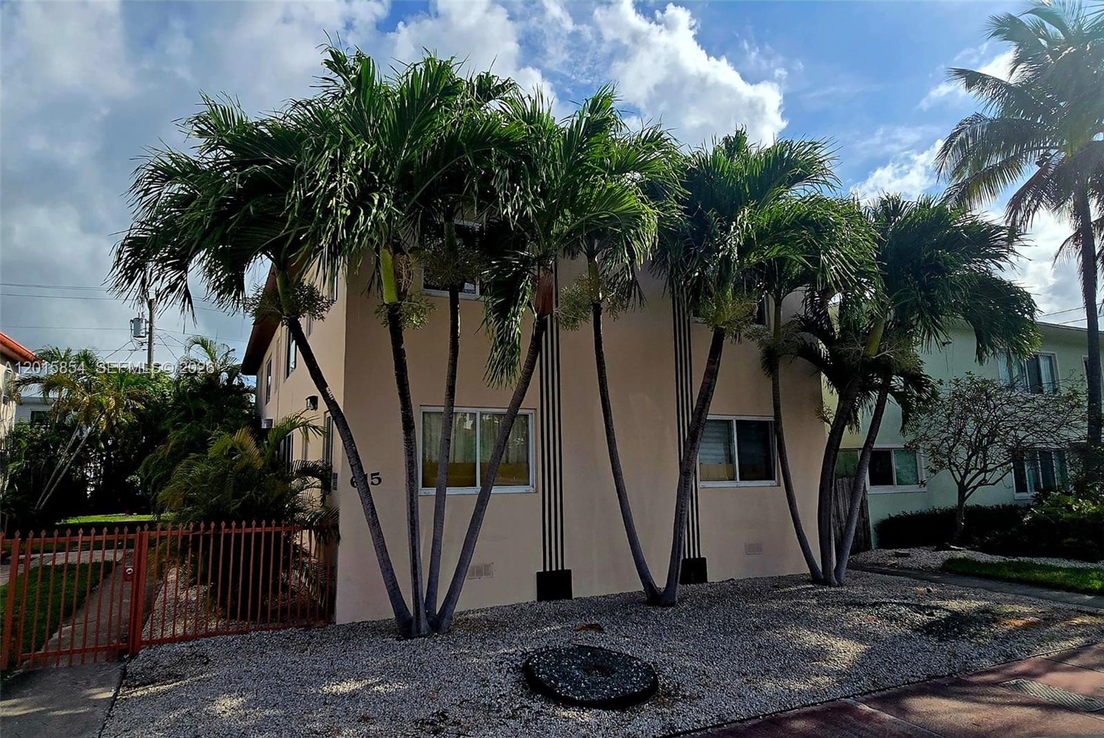 A two-story beige apartment building stands behind palm trees and a red metal fence — 615 82nd St # 1, Miami Beach FL 33141, MIAMI BEACH, FL