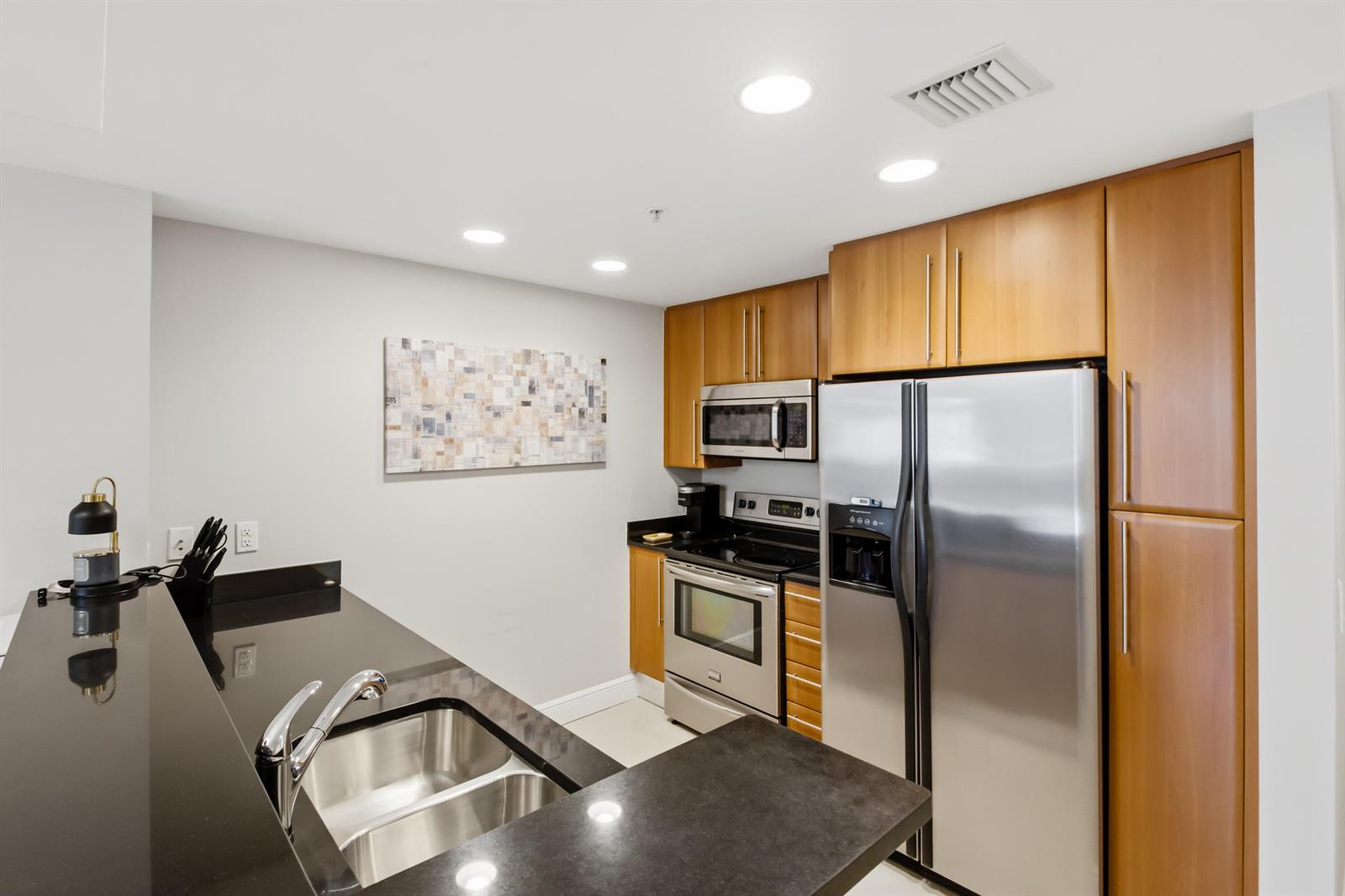 A kitchen features stainless steel appliances, wood cabinets, and black countertops with a double sink — 191 SE 20th Avenue # 210, Deerfield Beach FL 33441, DEERFIELD BEACH, FL