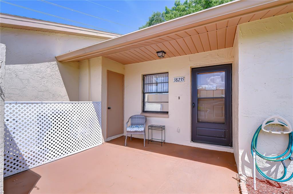 A covered patio features a brown door, a window, a chair, and a white lattice screen — 38235 BOXWOOD DRIVE UNIT 38235, ZEPHYRHILLS, FL