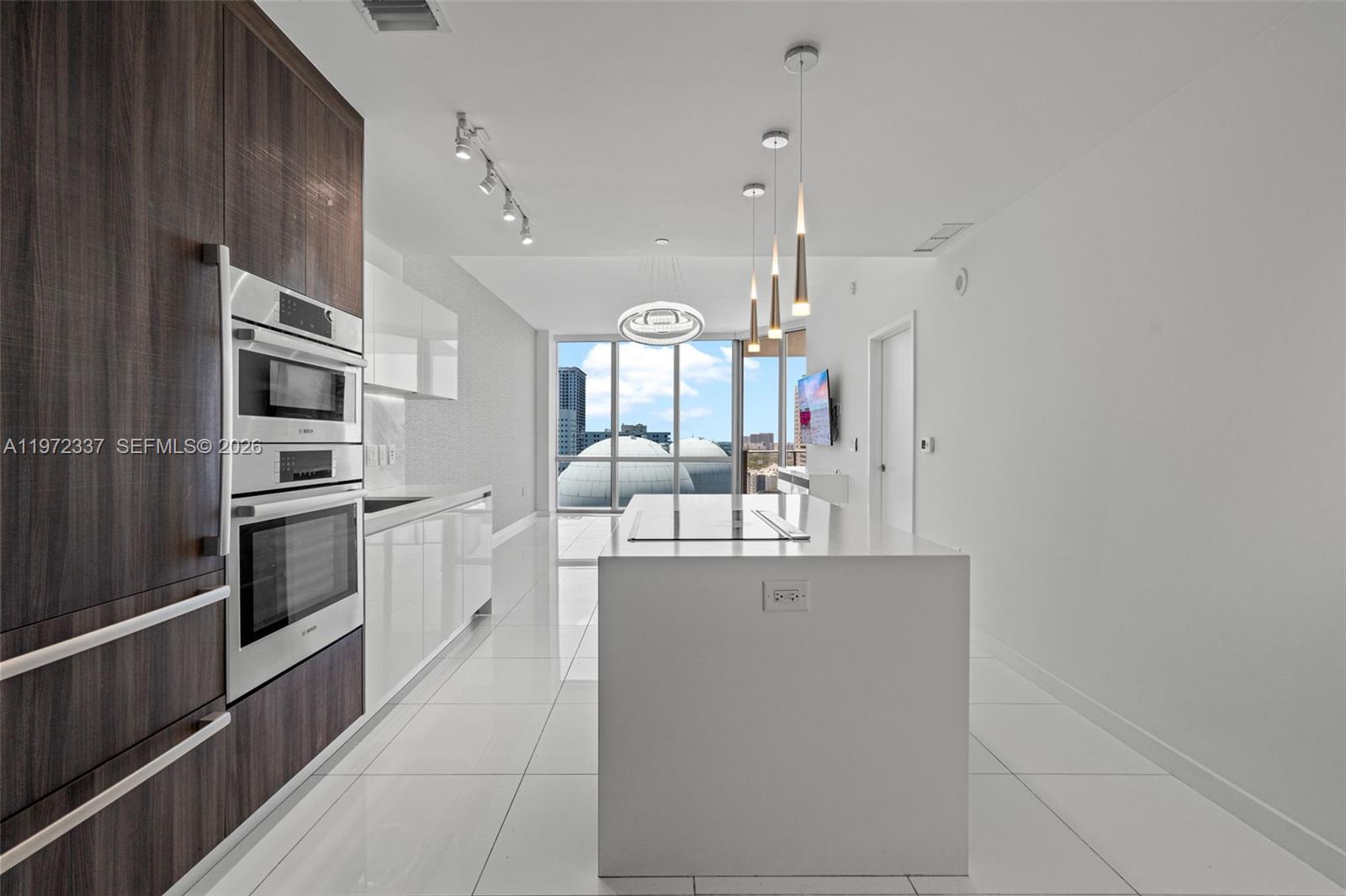 A modern kitchen features white cabinetry, a central island, and floor-to-ceiling windows with city views — 851 NE 1st Ave # 1506, Miami FL 33132, MIAMI, FL