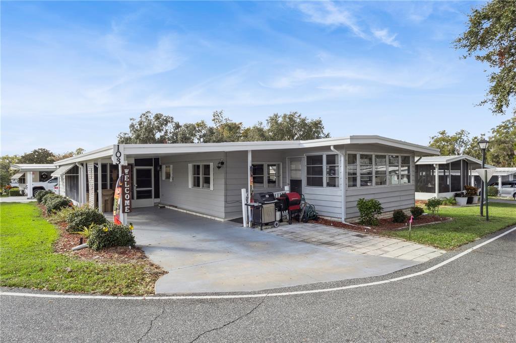 A single-story manufactured home with a gray exterior, carport, and a paved patio area — 103 POINSETTIA, LEESBURG, FL