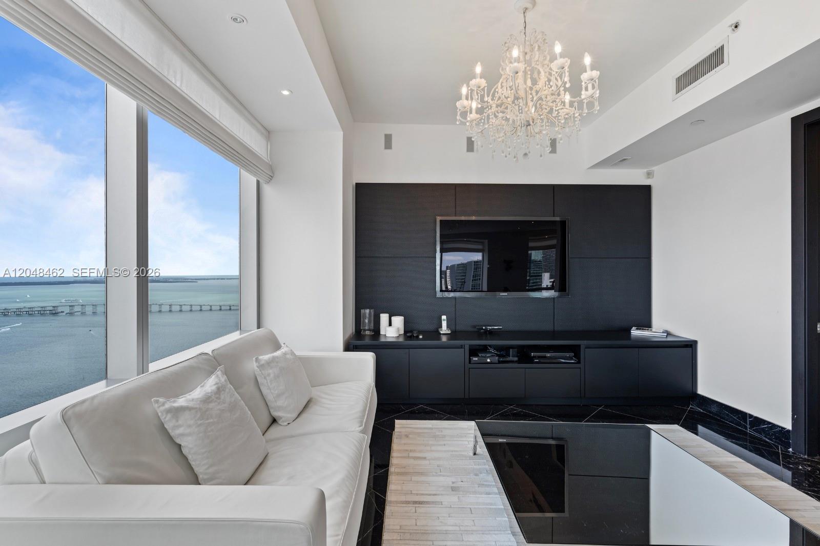 A modern living room features a white sofa, black media console, and a crystal chandelier — 1425 Brickell Ave # 42E, Miami FL 33131, MIAMI, FL