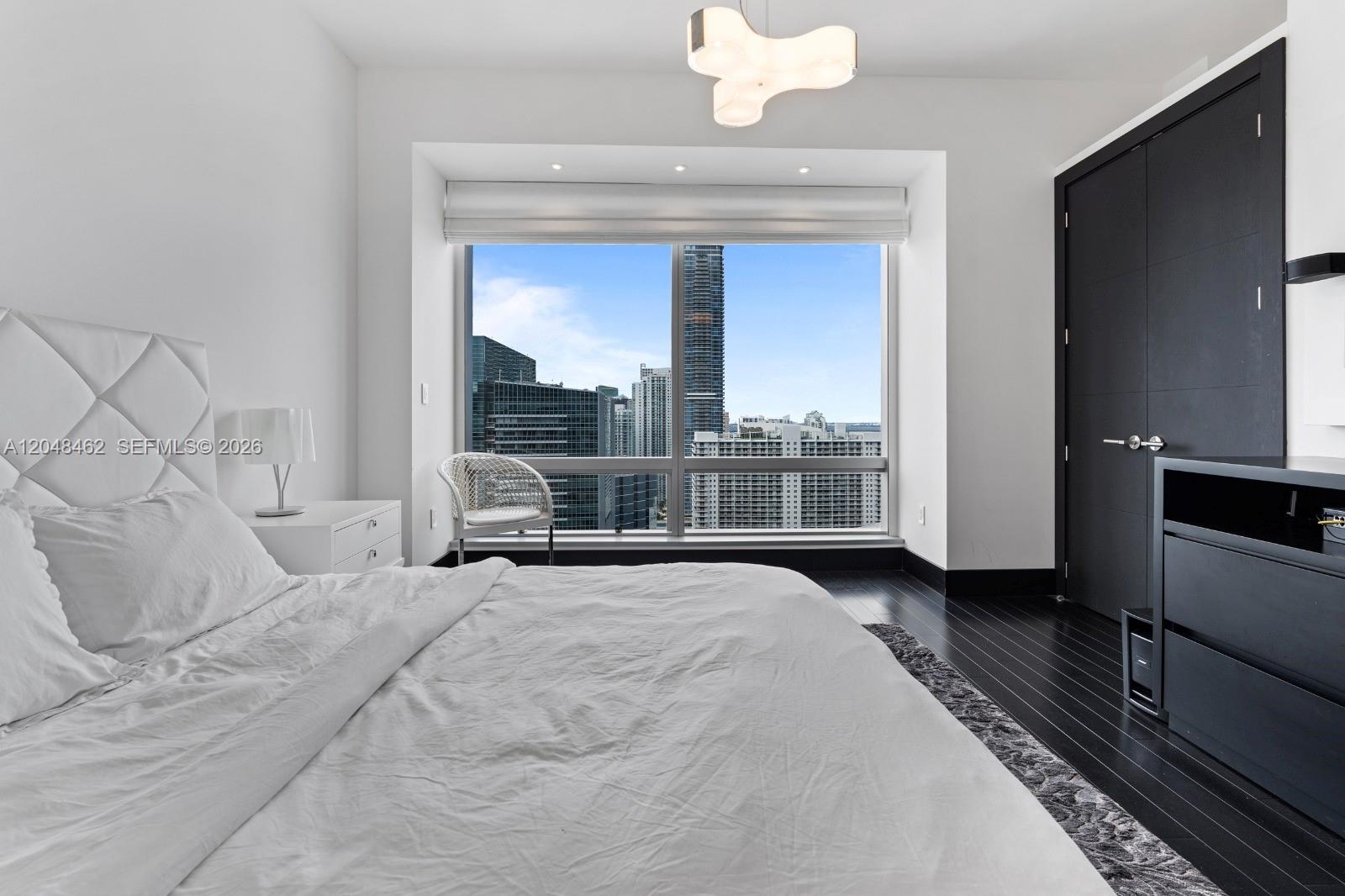 A modern bedroom features a white bed, dark wood floors, and floor-to-ceiling city view windows — 1425 Brickell Ave # 42E, Miami FL 33131, MIAMI, FL