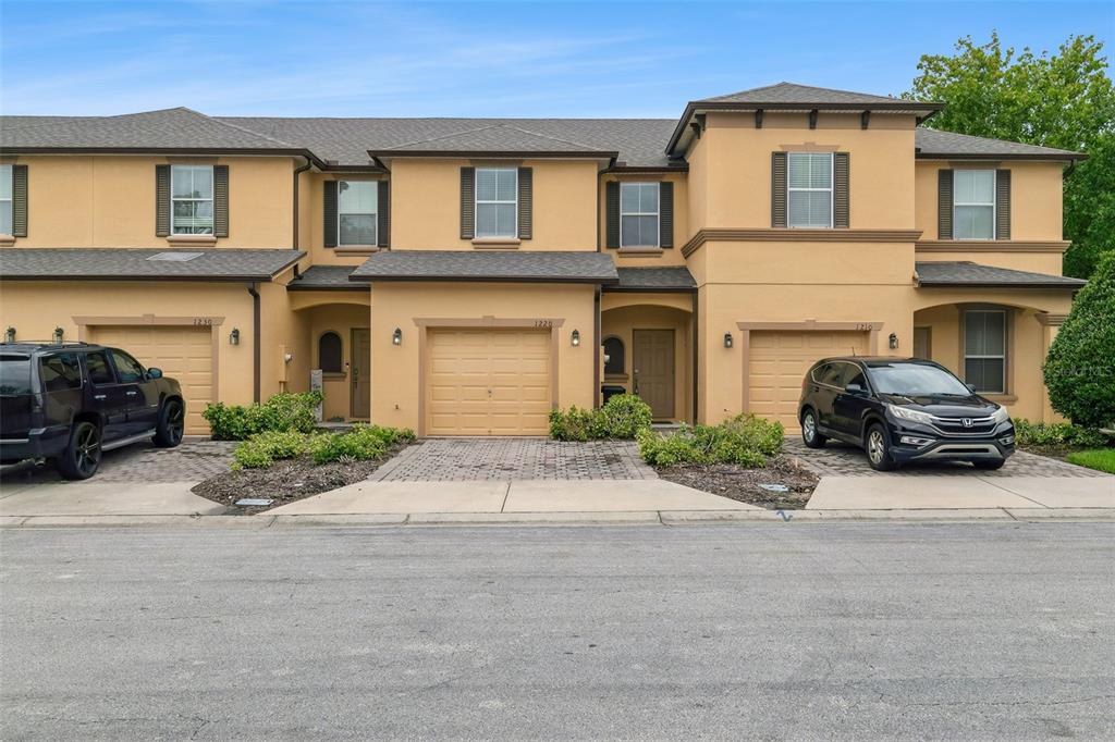 A row of three tan townhouses with attached garages and paved driveways under a blue sky — 1220 TWIN TREES LANE, SANFORD, FL