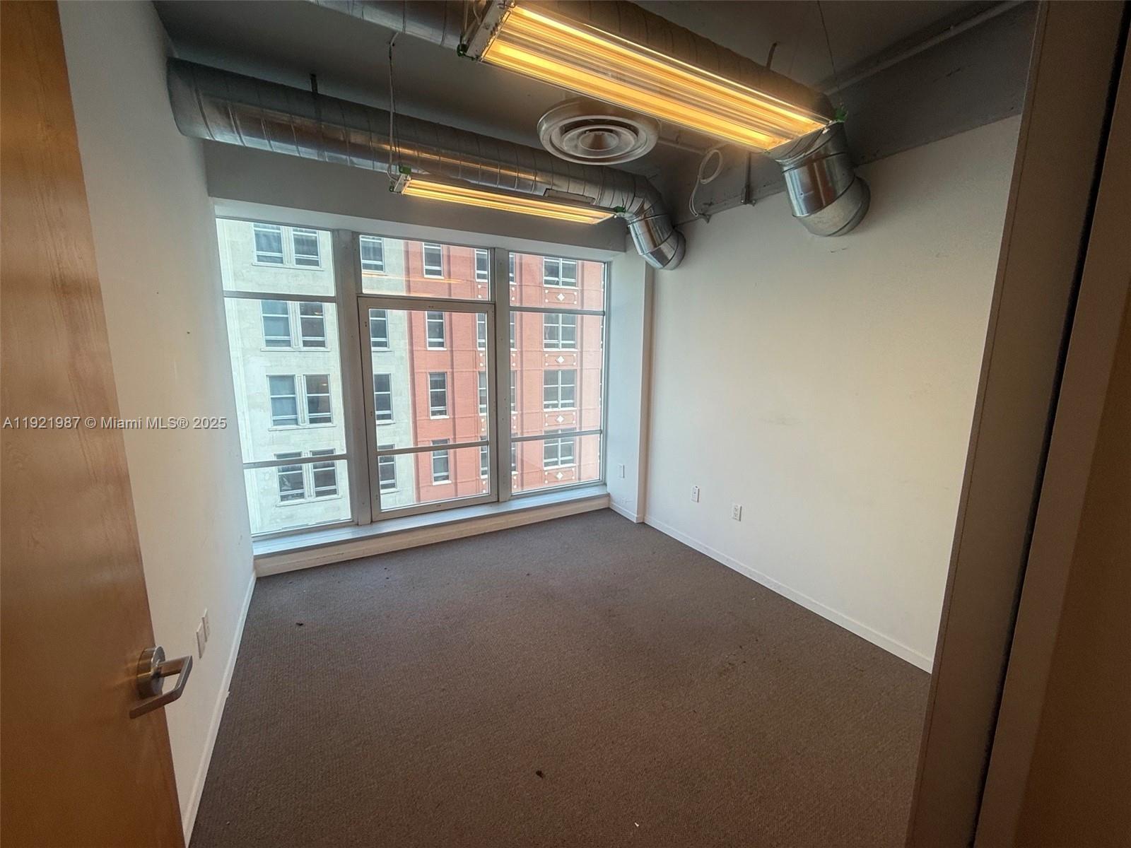 An empty office room features exposed ductwork, fluorescent lighting, carpeted flooring, and large windows — 14 NE 1st Ave # 800A, Miami FL 33132, MIAMI, FL