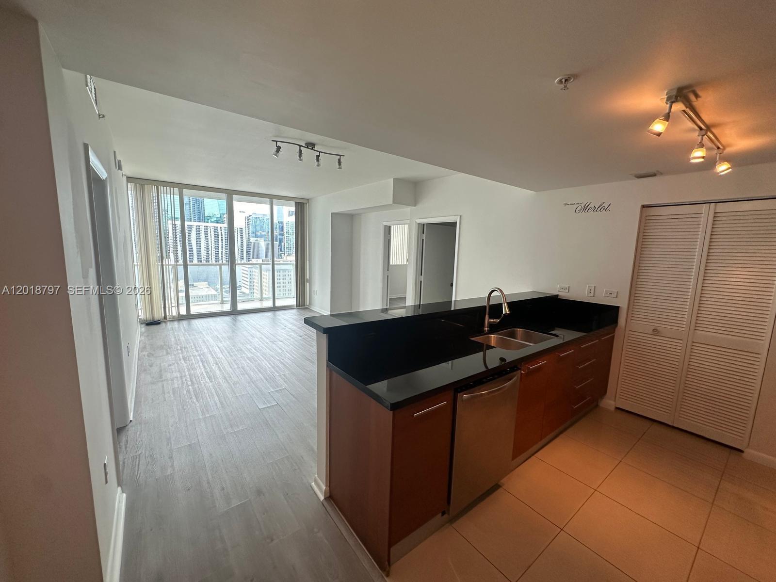 A kitchen with dark countertops and wood cabinets overlooks an open living area with balcony access — 244 Biscayne Blvd # 1910, Miami FL 33132, MIAMI, FL
