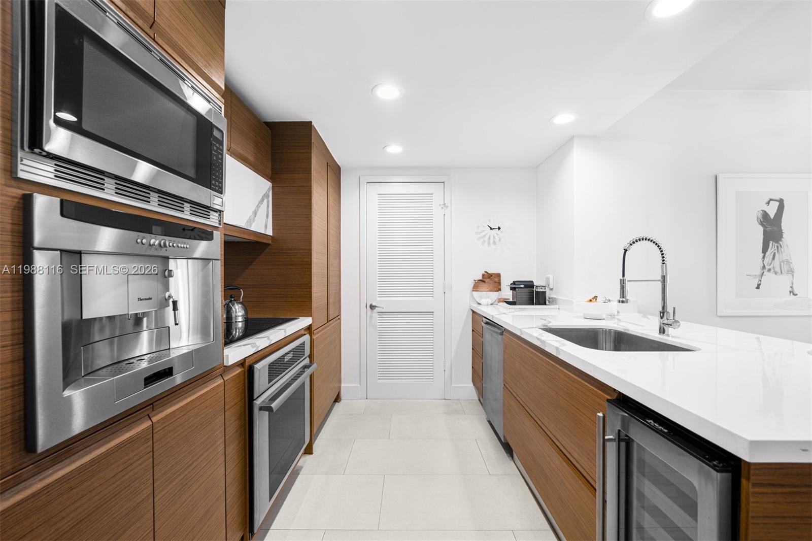 A modern galley kitchen features wood cabinetry, white countertops, stainless steel appliances, and tile flooring — 5875 Collins Ave # 906, Miami Beach FL 33140, MIAMI BEACH, FL