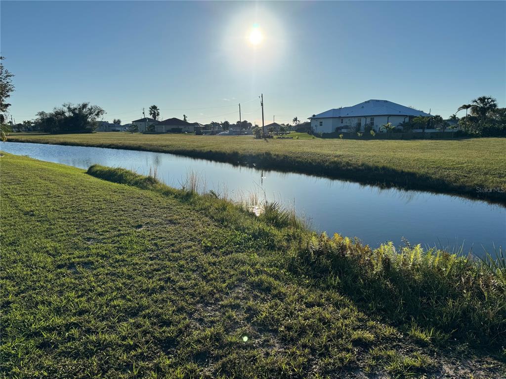 A grassy field borders a narrow canal under a bright sun with houses nearby — 17245 CAPE HORN BOULEVARD, PUNTA GORDA, FL