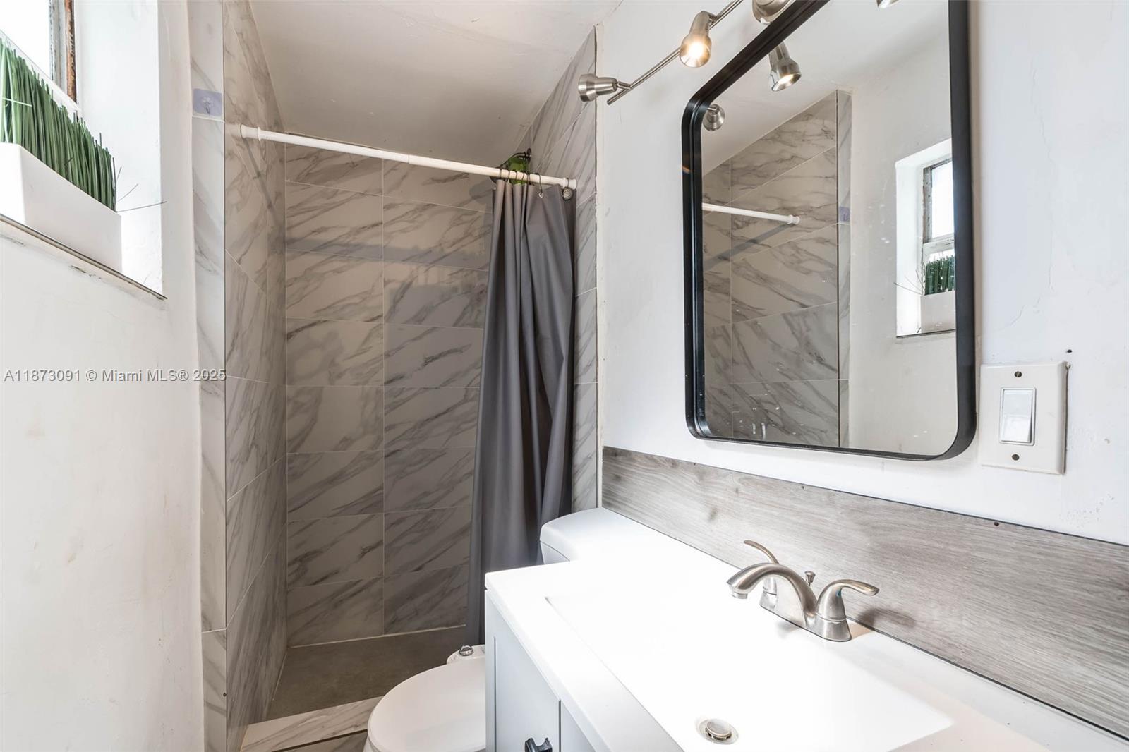 A bathroom features a marble-tiled shower, a white vanity, and a black-framed mirror — 180 NW 59th St, Miami FL 33127, MIAMI, FL