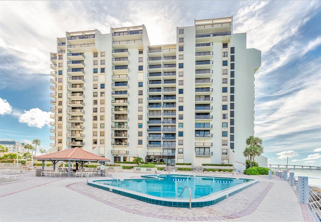 A multi-story residential building stands behind a swimming pool and a covered patio area — 440 S GULFVIEW BOULEVARD UNIT 1705, CLEARWATER BEACH, FL