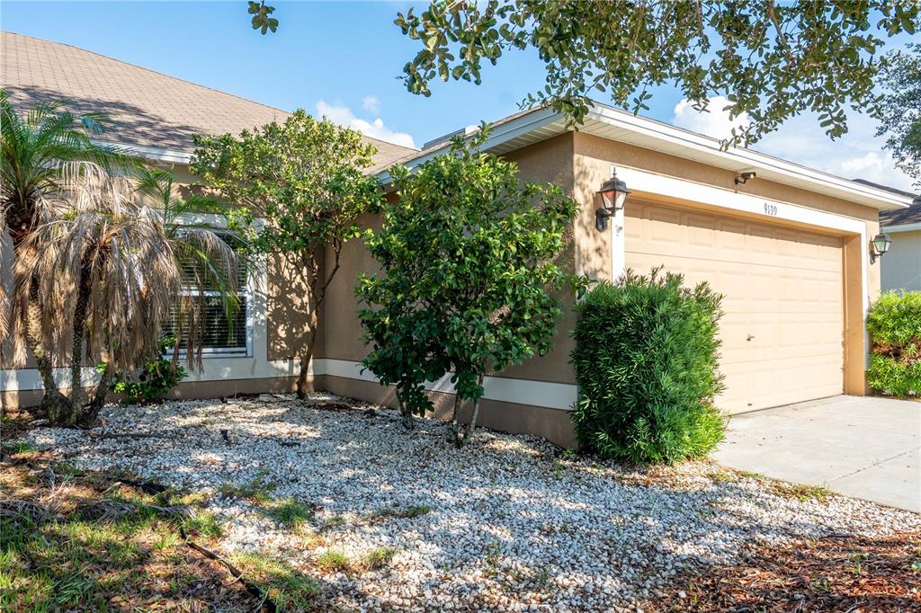 The exterior of a house features a tan stucco facade, a garage, and landscaped bushes — 9139 SOUTHERN CHARM CIRCLE, BROOKSVILLE, FL