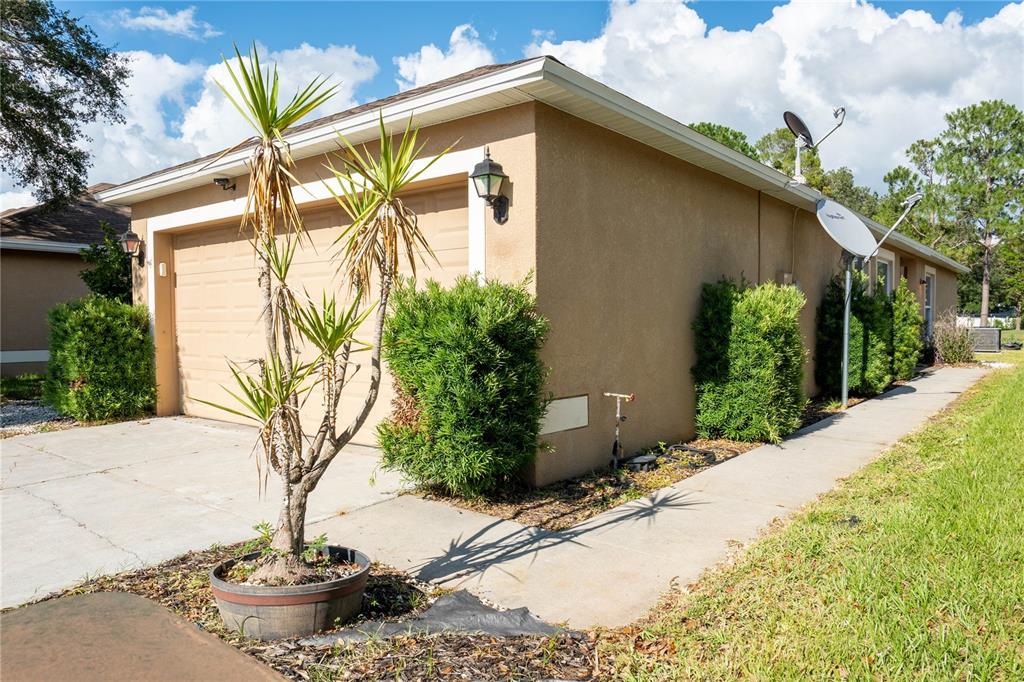 A tan stucco house exterior features a two-car garage, a potted palm, and landscaping — 9139 SOUTHERN CHARM CIRCLE, BROOKSVILLE, FL