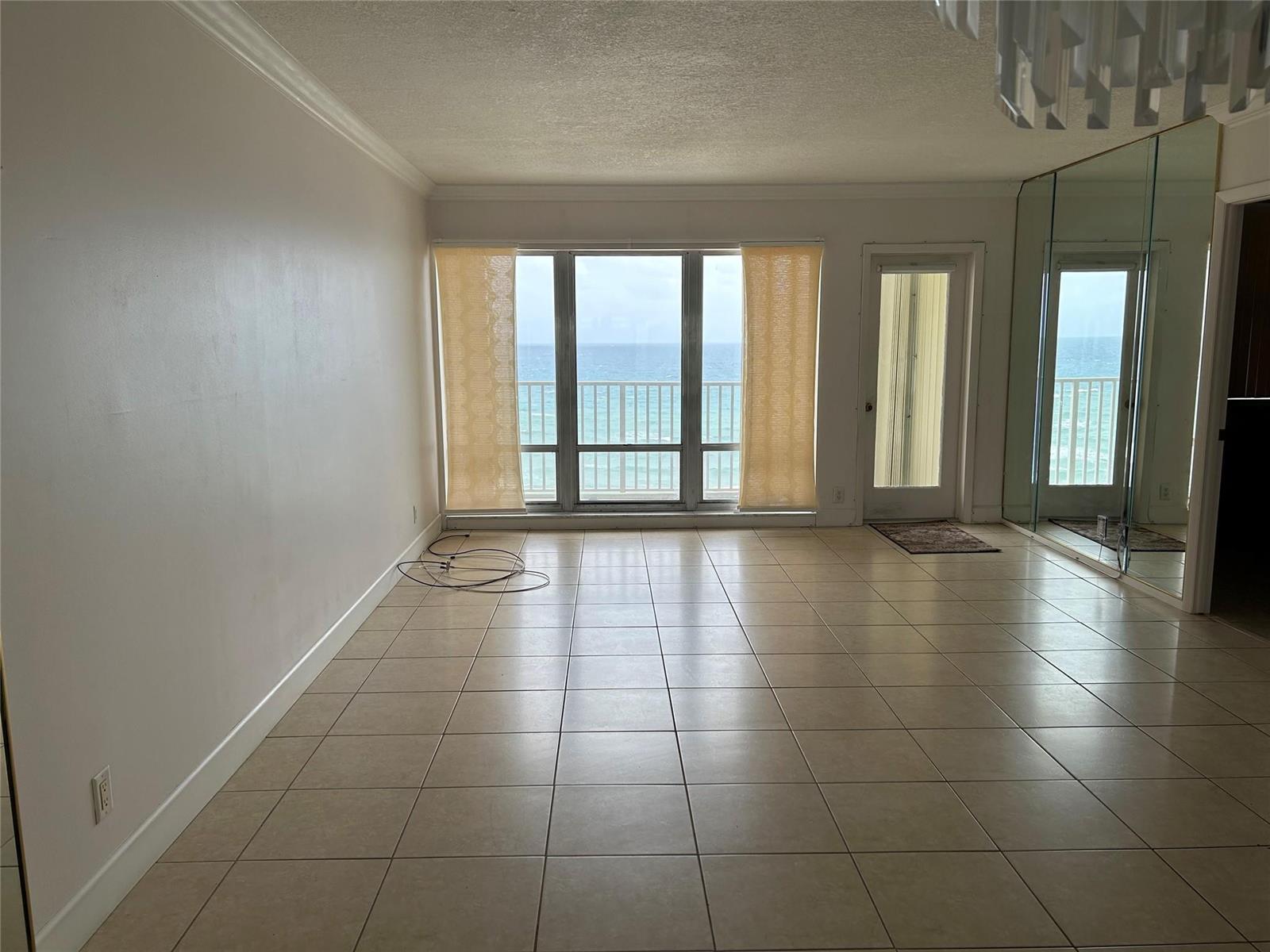 An empty living room features beige tile flooring, a large window, and a balcony door — 750 N Ocean Boulevard # 1409, Pompano Beach FL 33062, POMPANO BEACH, FL