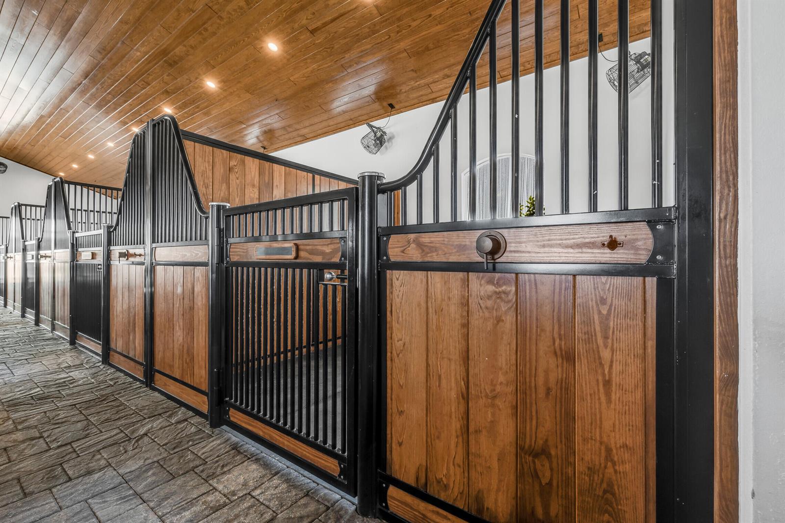 A row of horse stalls features wood-paneled walls, black metal grilles, and stone flooring — 5181 Lasso Way, Wellington FL 33449, WELLINGTON, FL